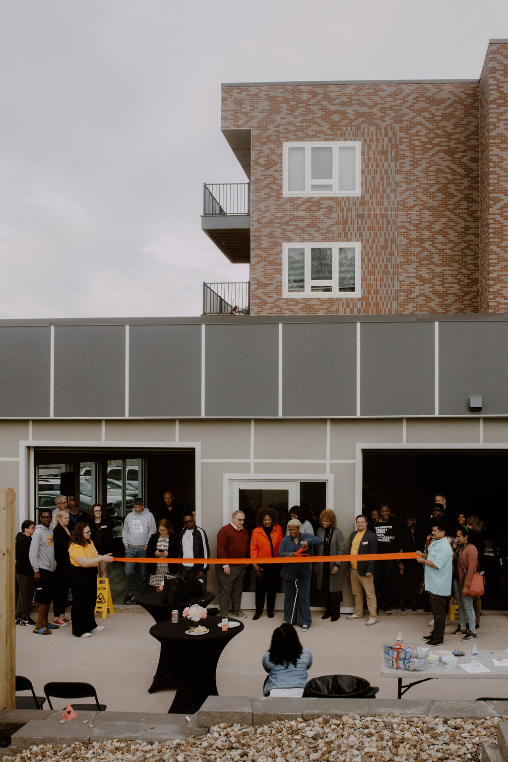 A group of people stand behind an orange ribbon in front of a brand new housing development.