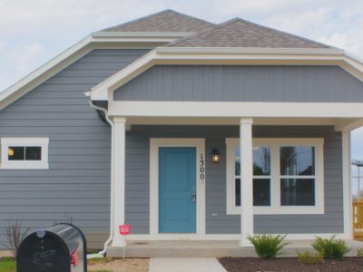 A one-story house with a door and two windows on the front. The home is painted a blueish gray and the door is a turquoise blue.