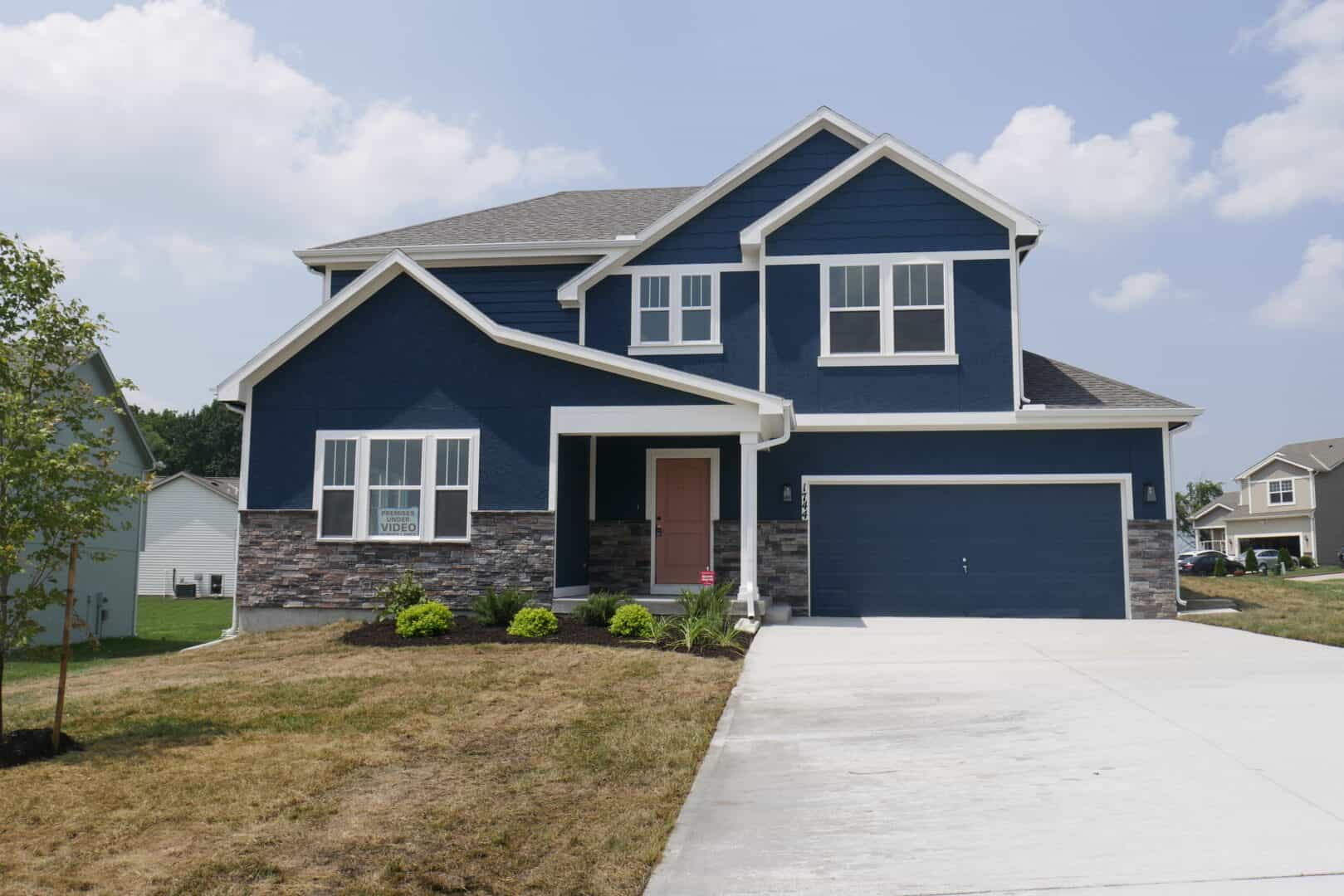 A two-story home with an attached garage and landscaping out front. The house is painted a rich dark blue with white trim. Three windows are visible on the front face of the home, one on the first floor and two on the second floor. The front entry is visible; the door is painted a light brown or red.