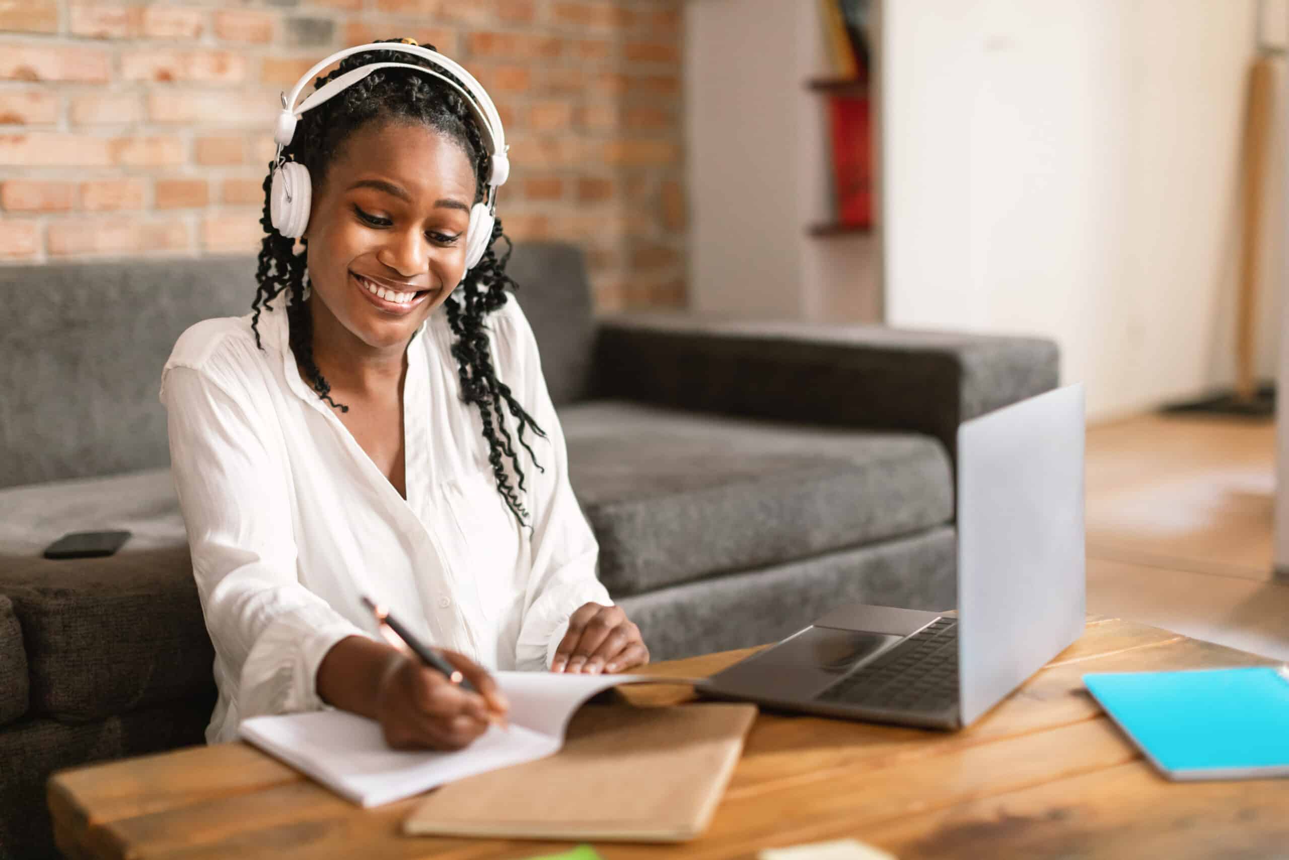 Happy african american woman studying online from home, taking notes, sitting in front of laptop wearing headphones, copy space. Modern distance education and technology