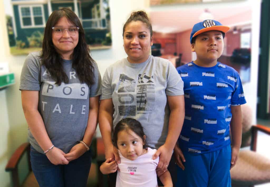 Rita and her family stand together in front of a photo of a beautiful home. They all have brown skin and dark brown hair. Rita's daughter and son stand next to her on either side with friendly expressions. She holds her younger daughter in her arms in front of her.
