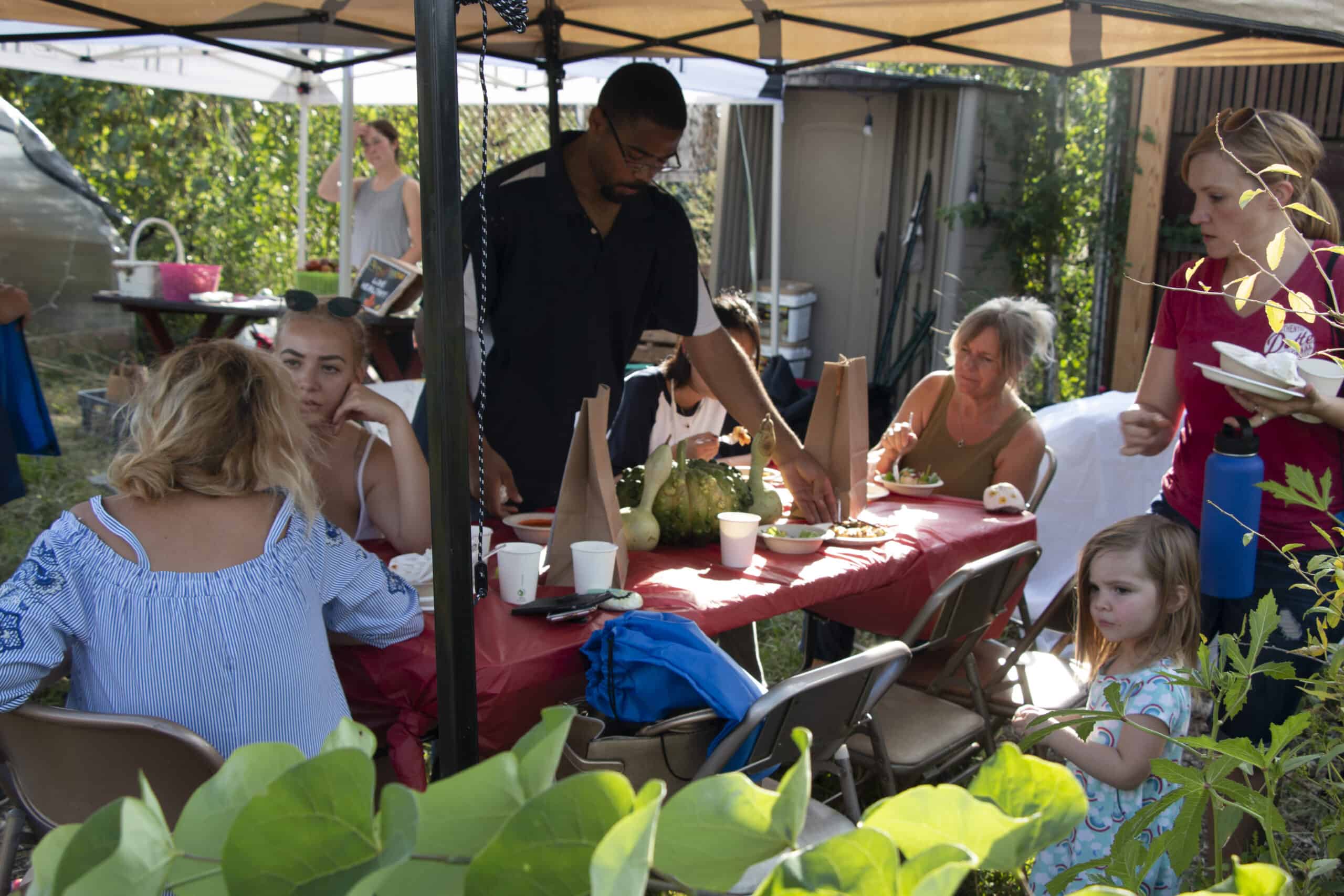 A group of families sit down at a table together eating a freshly prepared meal at a picnic table outside.