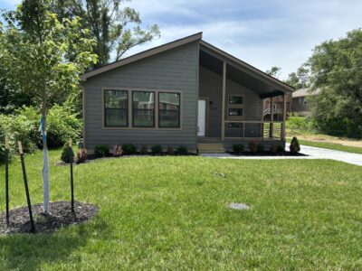 A single-story home with green paint and brown trim, surrounded by a driveway, green grass and blue sky.