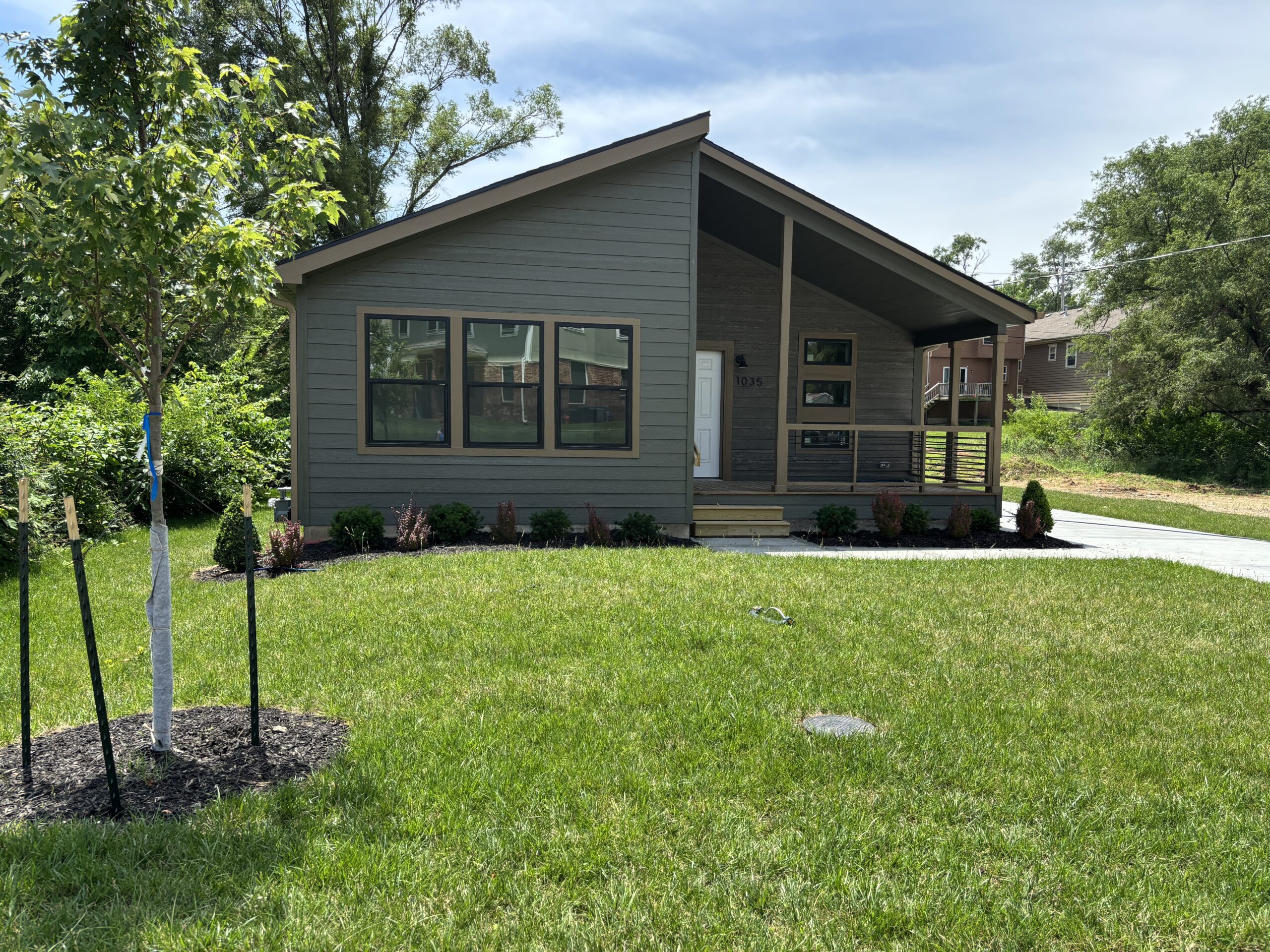 A single-story home with green paint and brown trim, surrounded by a driveway, green grass and blue sky.