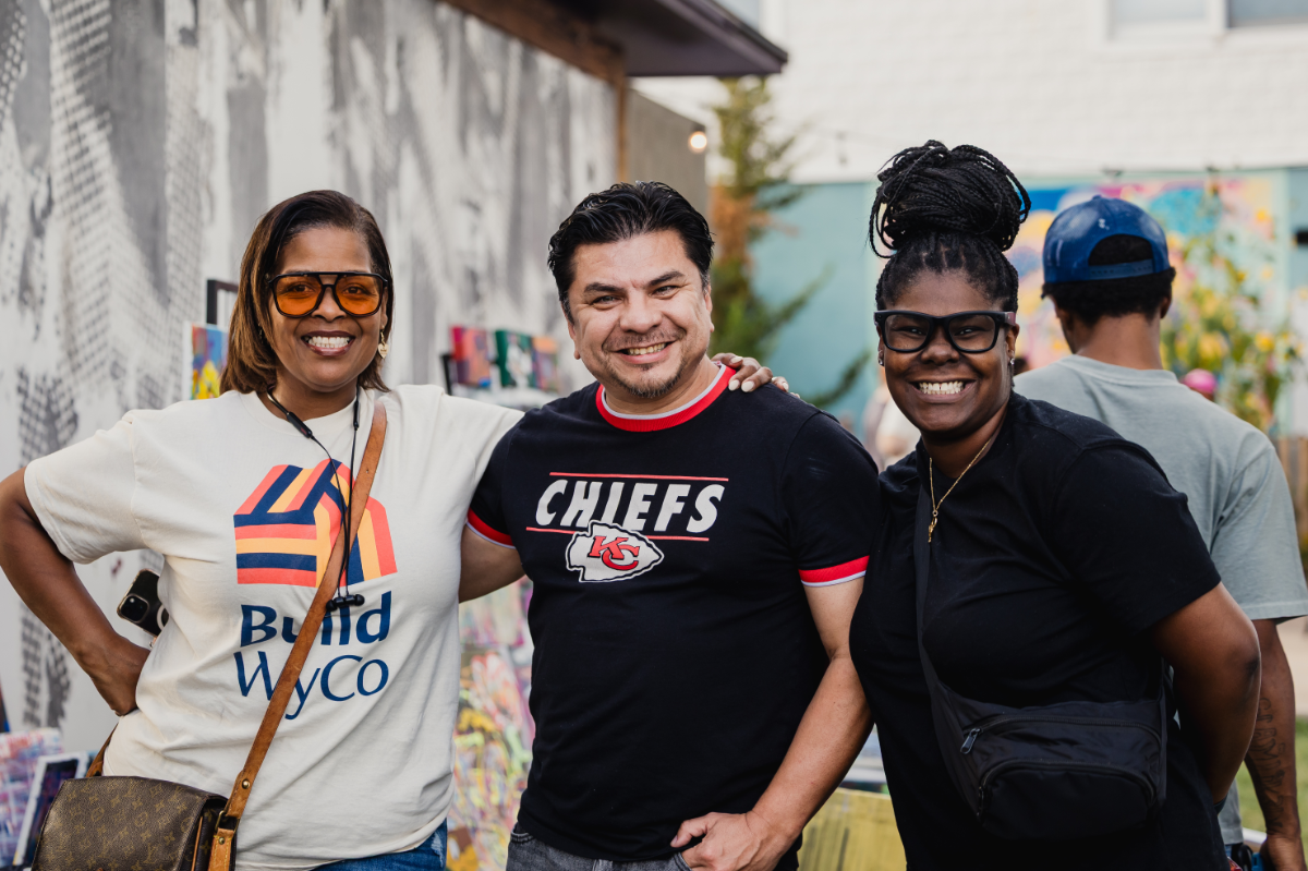 A photo of three people standing together at an outdoor event. They are smiling and wearing Build WyCo and Chiefs shirts.