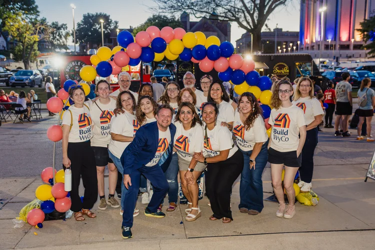 A group of people wearing a house with a colorful house logo stand in front of a balloon arch outside.
