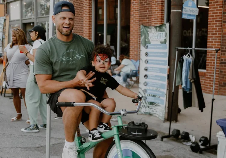 A man and a boy ride a bike at an outdoor festival outside a storefront.