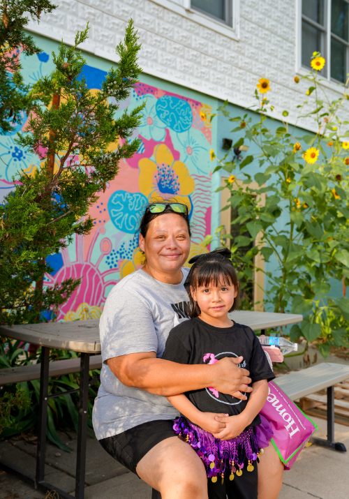 a woman and child sit outside next to a colorful floral mural and some greenery.