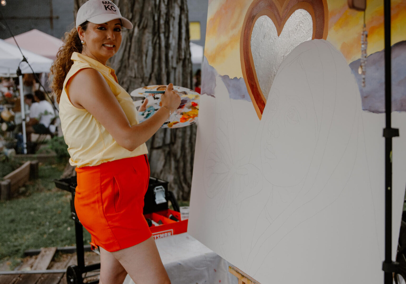 A woman stands in front of a partially painted canvas holding a palette and brush.